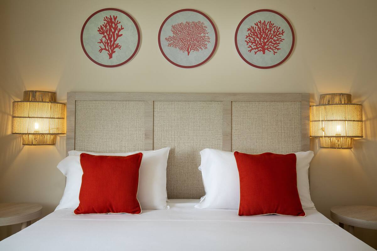 A hotel room with a bed, two red and white pillows, a headboard, and three wall-mounted lamps.