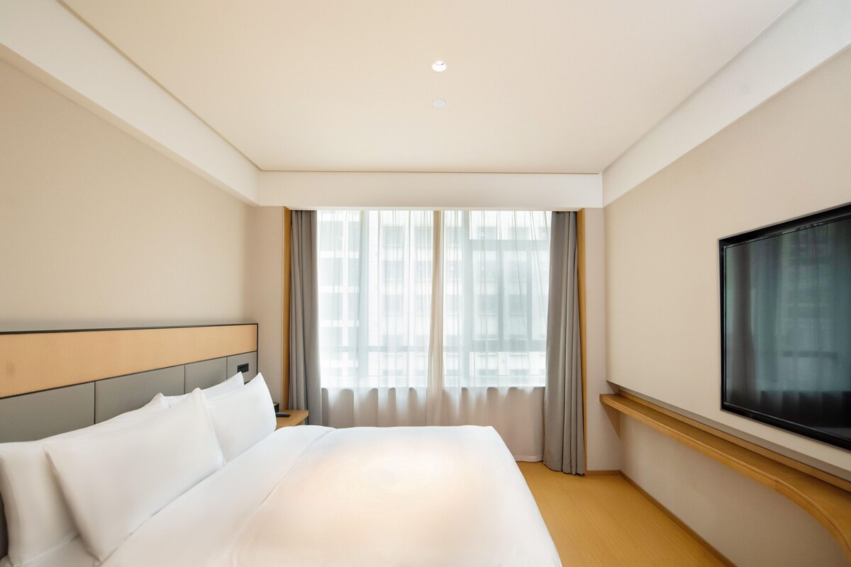 A hotel room with a large bed, a television on the wall, and a window with curtains.