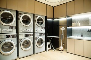 A laundry facility with multiple washing machines and dryers, a ironing board, and a vacuum cleaner. - Ji Hotel (Chongqing)