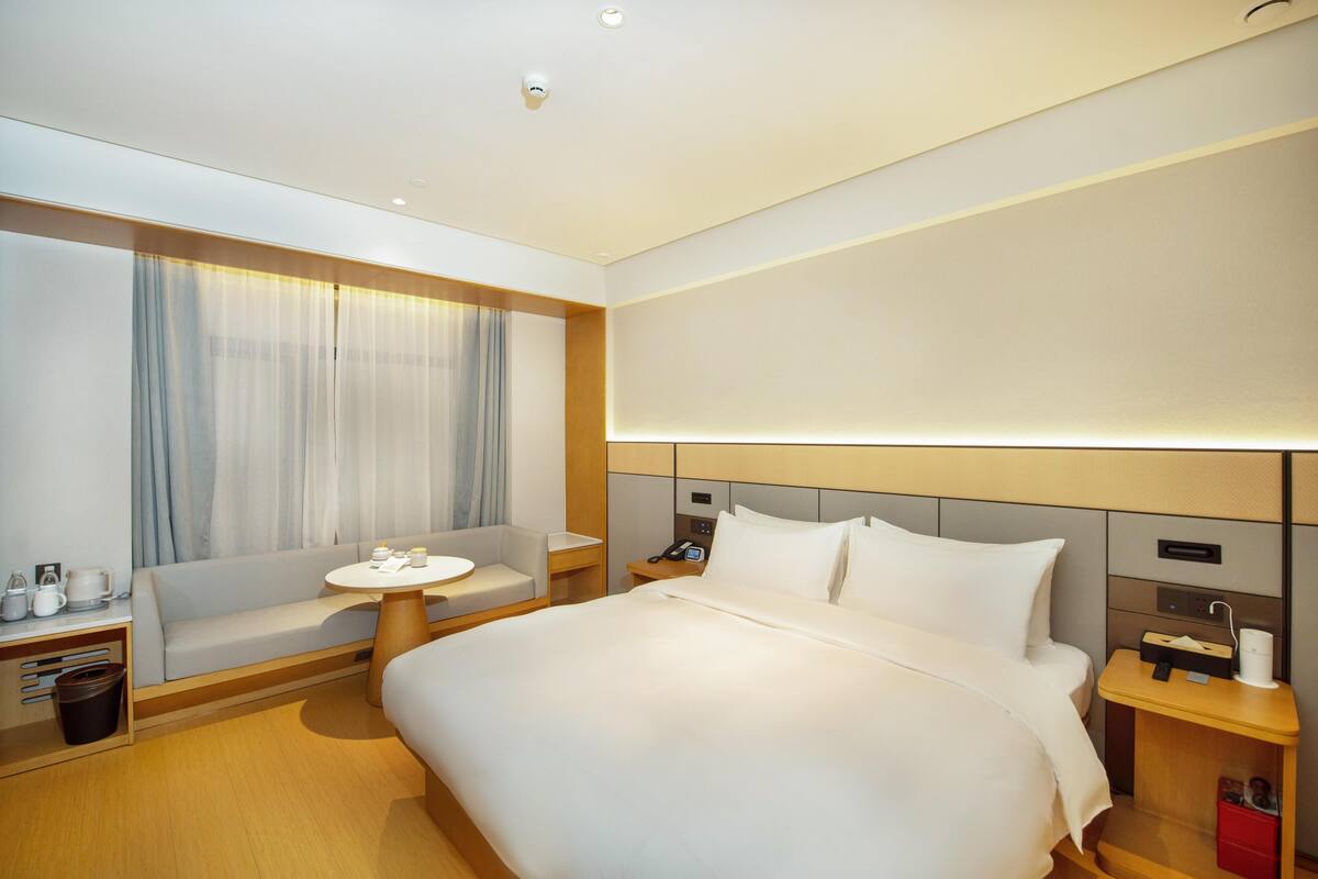 A modern hotel room with a large bed, a sofa, a small table, and a nightstand with a phone and a lamp.