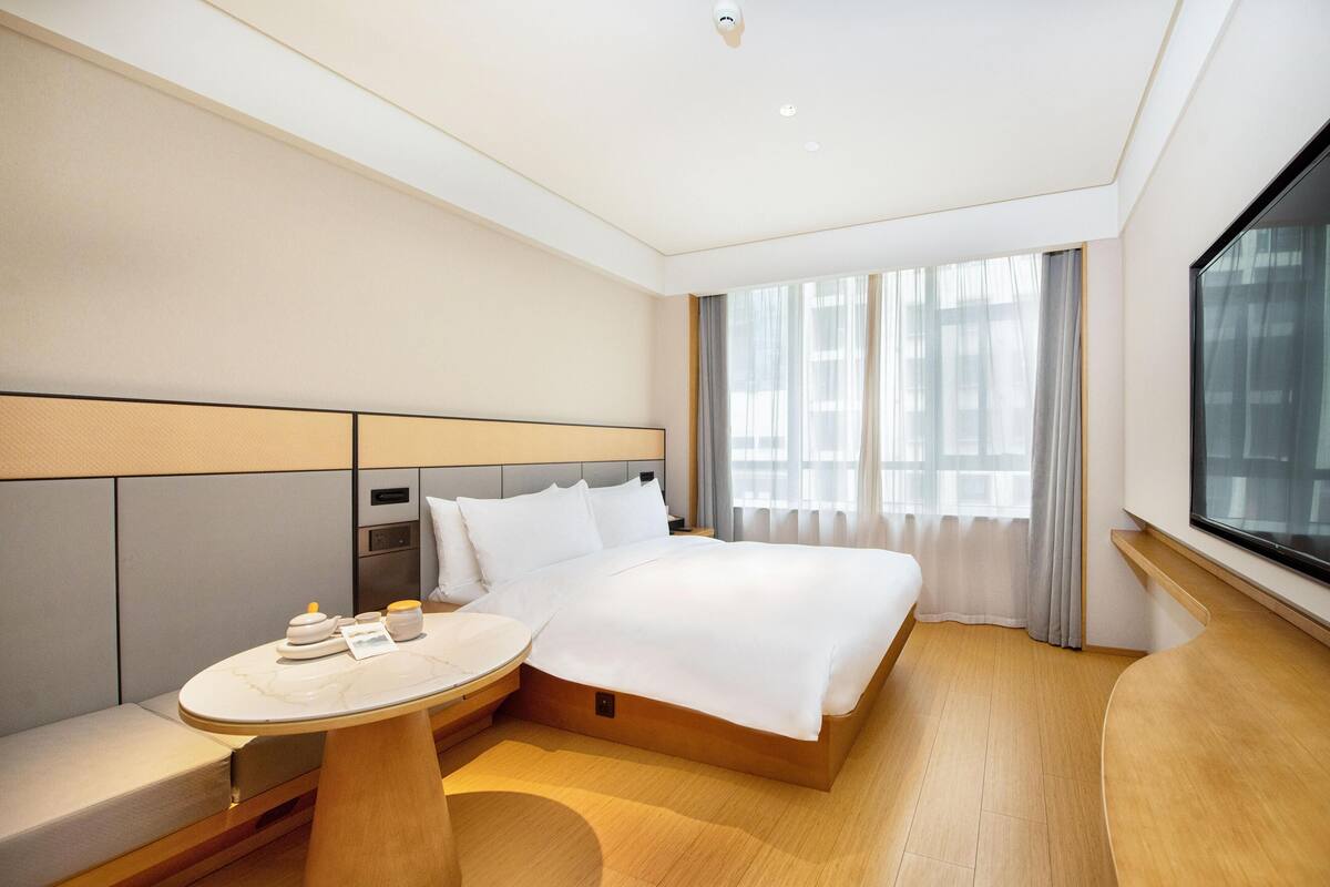 A modern hotel room with a large bed, bedside table, and a view of the city through large windows.