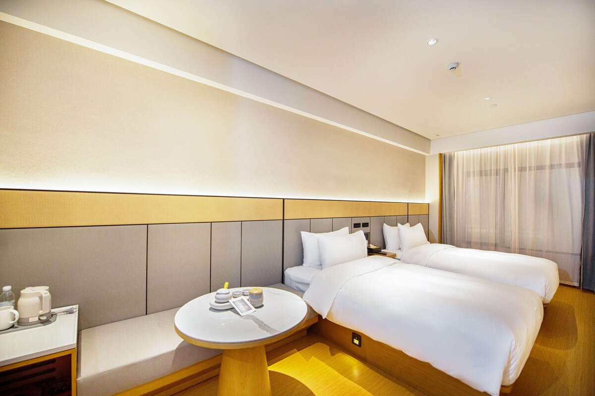 A hotel room with two beds, a small round table with a coffee maker, and a window with curtains.