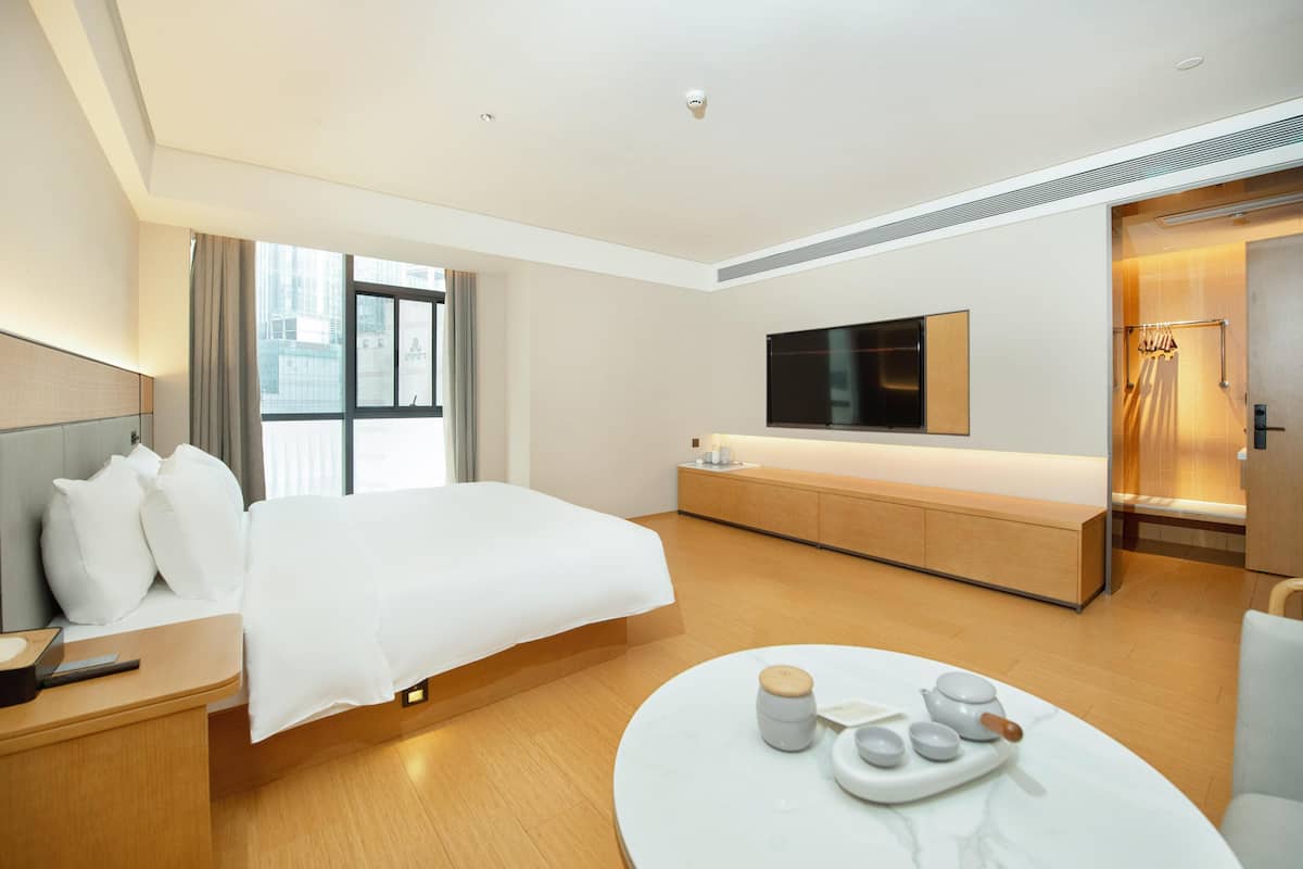 A modern hotel room with a large bed, a bedside table, a round coffee table with tea set, a TV mounted on the wall, and a view of the city through the window.