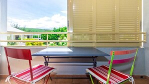 A balcony with a table and two chairs, a window with blinds, and a view of greenery. - Purple - Cosy duplex in the heart of L'Hermitage, close to the lagoon (Saint-Paul)