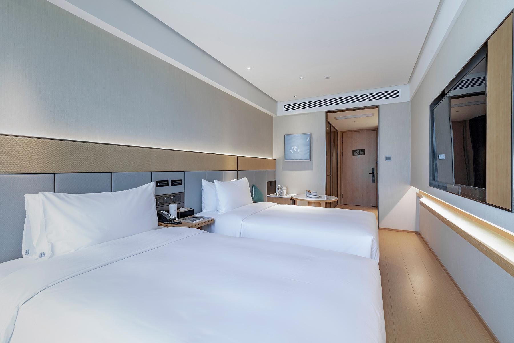 A hotel room with two beds, a bedside table with a lamp, a small round table, and a large mirror.
