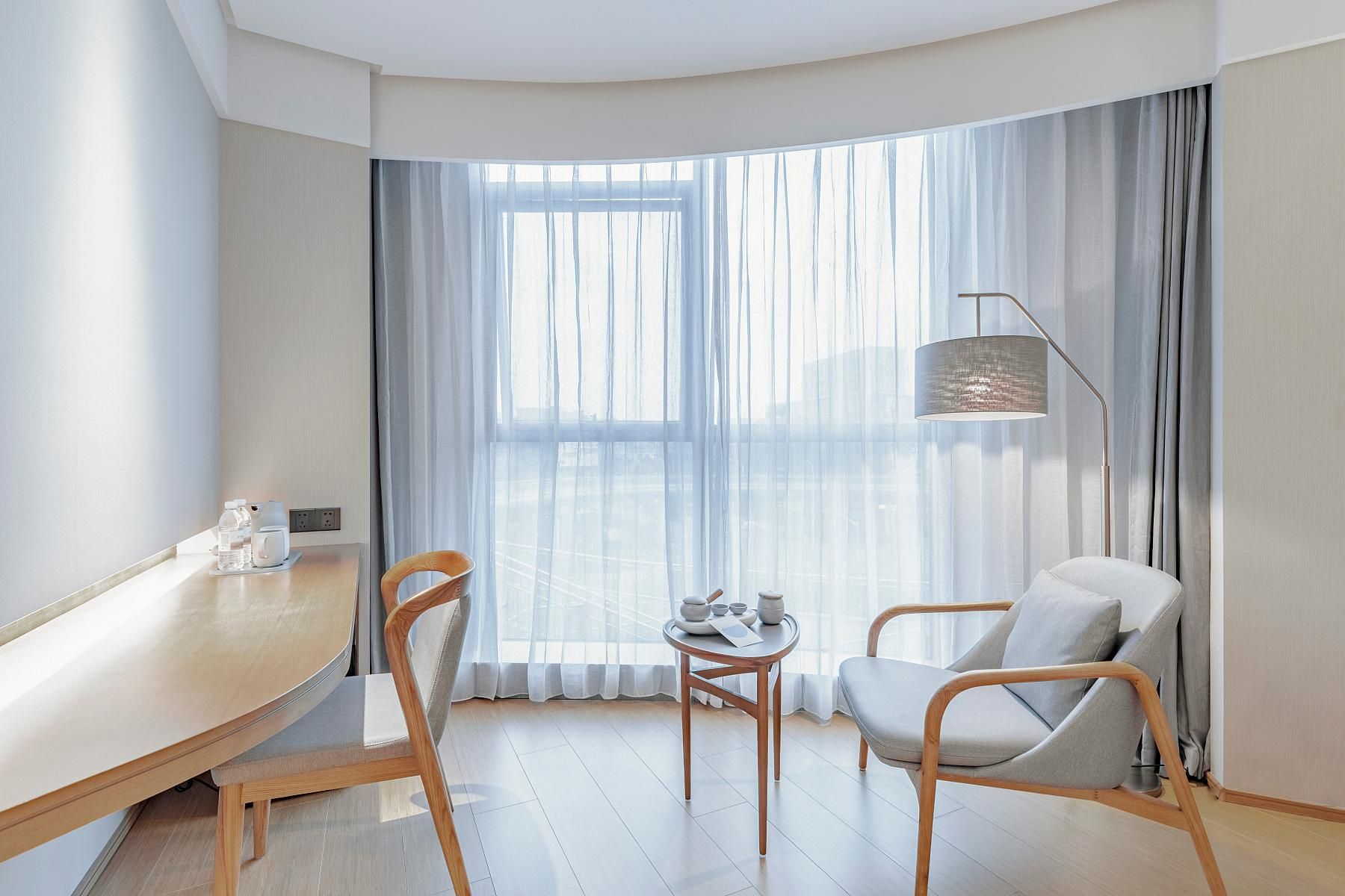 A modern hotel room with a curved ceiling, a large window with sheer curtains, a wooden desk with a chair, a round table with a chair, and a floor lamp.