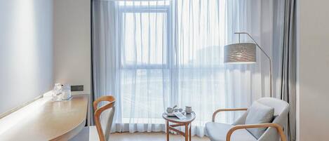 A modern hotel room with a curved ceiling, a large window with sheer curtains, a wooden desk with a chair, a round table with a chair, and a floor lamp.