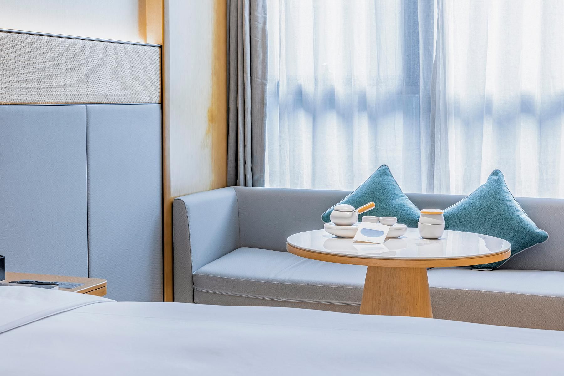 A hotel room with a bed, a round table with tea set, a sofa with cushions, and a window with curtains.