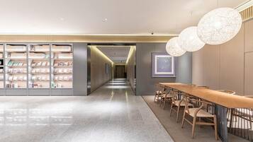 A modern hotel lobby with a long reception desk, a bookshelf with magazines, and a hallway leading to the dining area.
