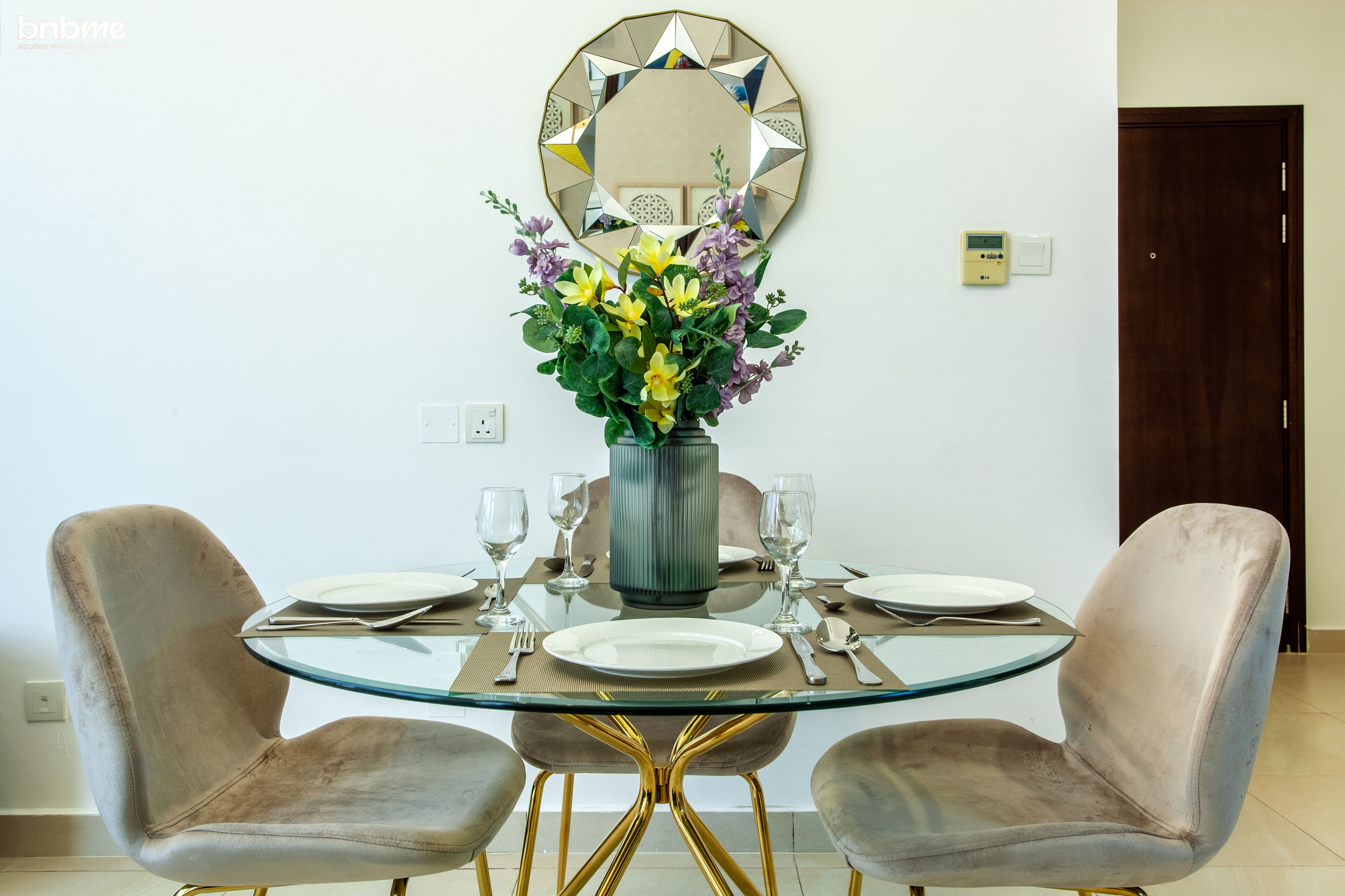 A dining area with a round glass table, two cushioned chairs, a vase with flowers, and a decorative mirror on the wall.