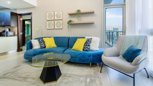 Un salon moderne avec un canapé bleu, une table basse en verre et un fauteuil gris. Des étagères murales accueillent des plantes et des œuvres d’art encadrées.