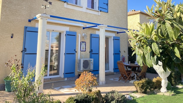 A two-story house with blue shutters, a covered patio with a table and chairs, and a garden with various plants. - Villa with terrace and garden near the beach (Narbonne plage)