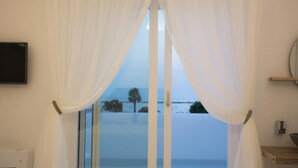 Una habitación con un ventanal grande cubierto por cortinas blancas, un televisor de pantalla plana montado en la pared y una estantería de madera con un cuenco.