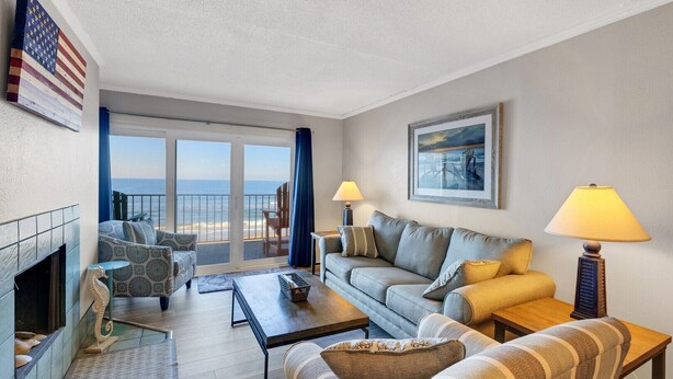 A living room with a fireplace, a sofa, a coffee table, and a view of the ocean through large windows. - DL403 Tranquility Shore by First Flight Rentals (Nags Head)