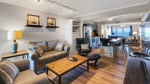 A modern living room with a sofa, coffee table, and dining area. There is a kitchen in the background with a refrigerator and stove. - DL403 Tranquility Shore by First Flight Rentals (Nags Head)
