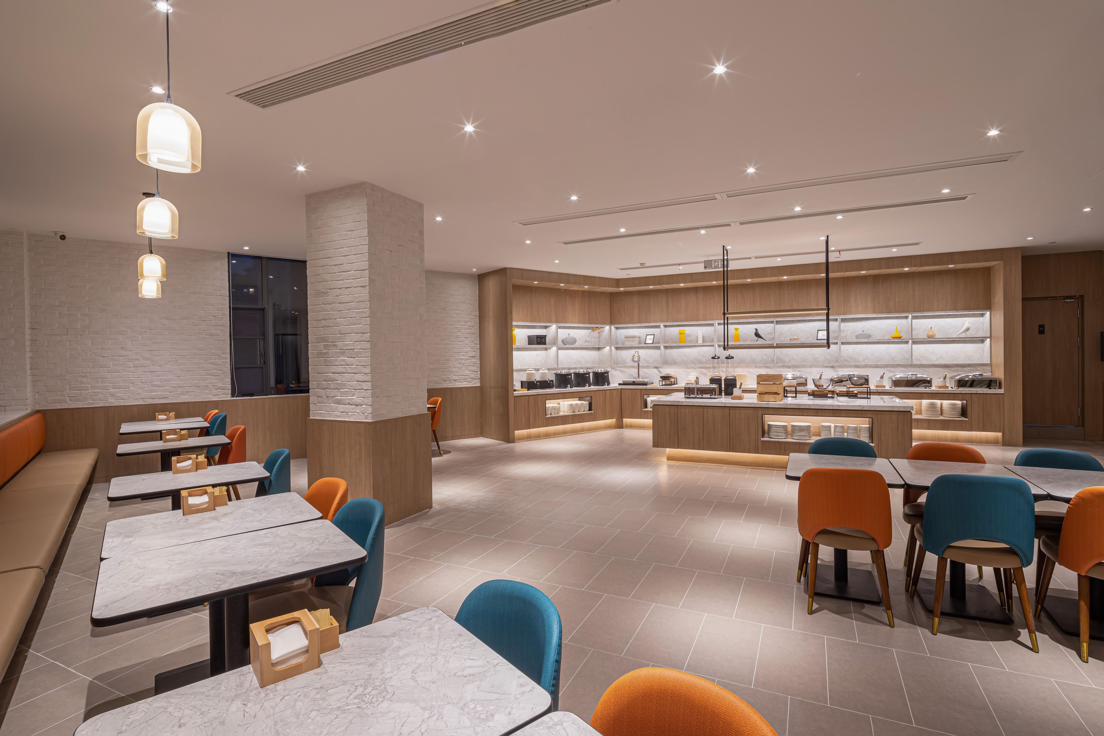 Een modern restaurant met marmeren tafels, oranje en blauwe stoelen, en een centraal gelegen keuken.