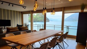 A modern dining area with a wooden table and chairs, a sofa with cushions, and a view of a lake and mountains through large windows. - Chalet Apartments Iseltwald (Iseltwald)