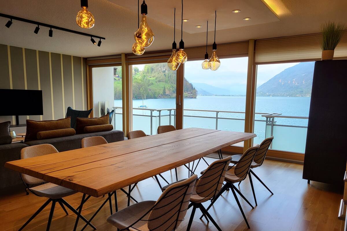 A modern dining area with a wooden table and chairs, a sofa with cushions, and a view of a lake and mountains through large windows.