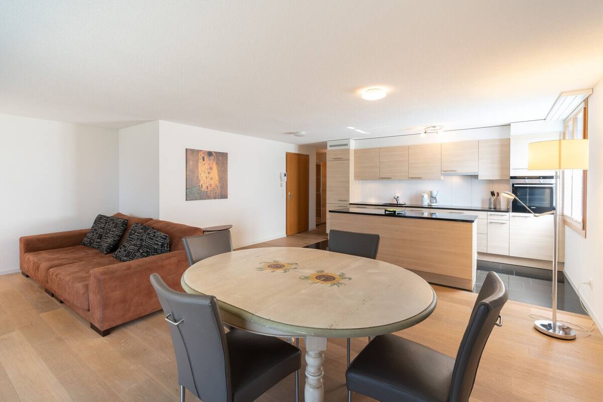 A modern living room with a brown sofa, a round table with chairs, and a kitchen in the background.