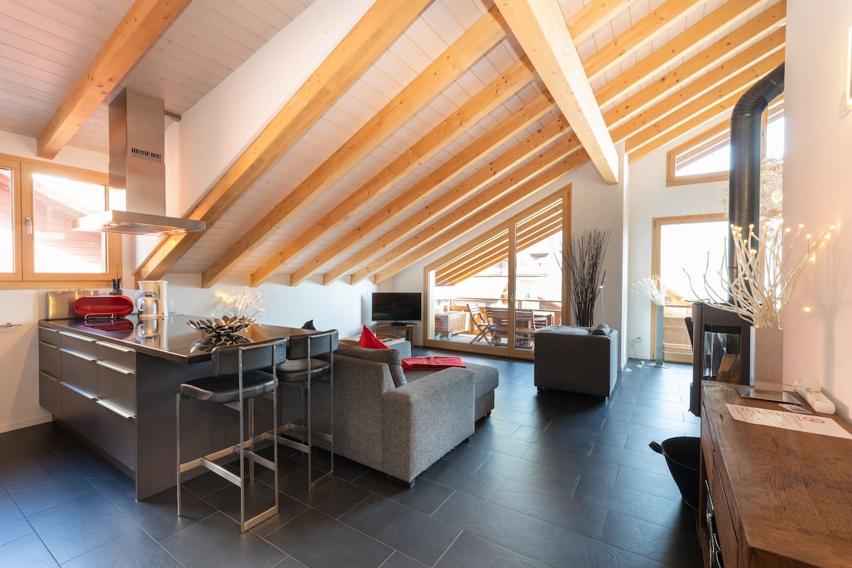 A modern loft-style living space with a high ceiling, built-in kitchen, and large windows.
