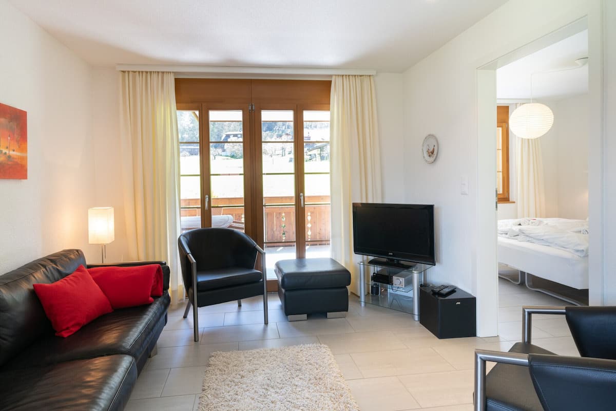 A modern living room with a leather sofa, a black armchair, a flat-screen TV, and a balcony with curtains.