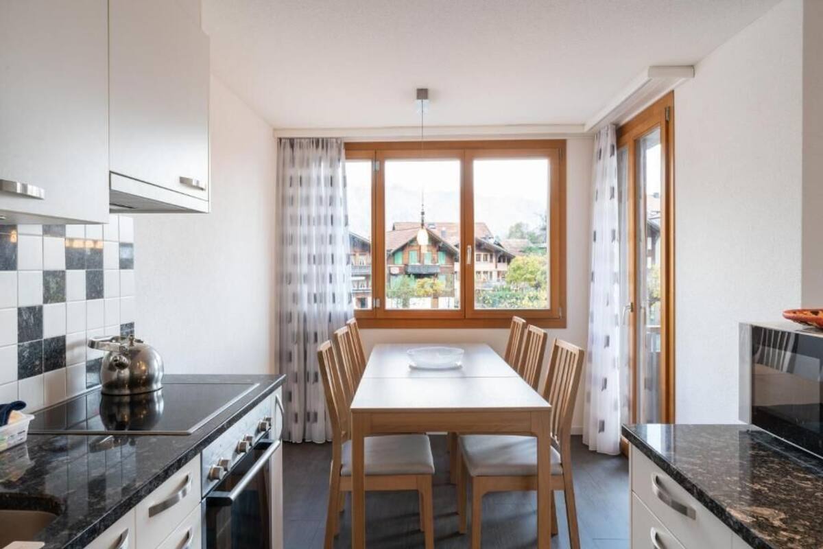 A modern kitchen with a dining table, wooden chairs, and a view of a mountain village through large windows.