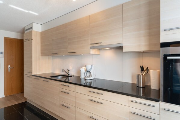 Una cocina moderna con armarios de madera clara, encimeras negras, electrodomésticos de acero inoxidable y una cafetera.