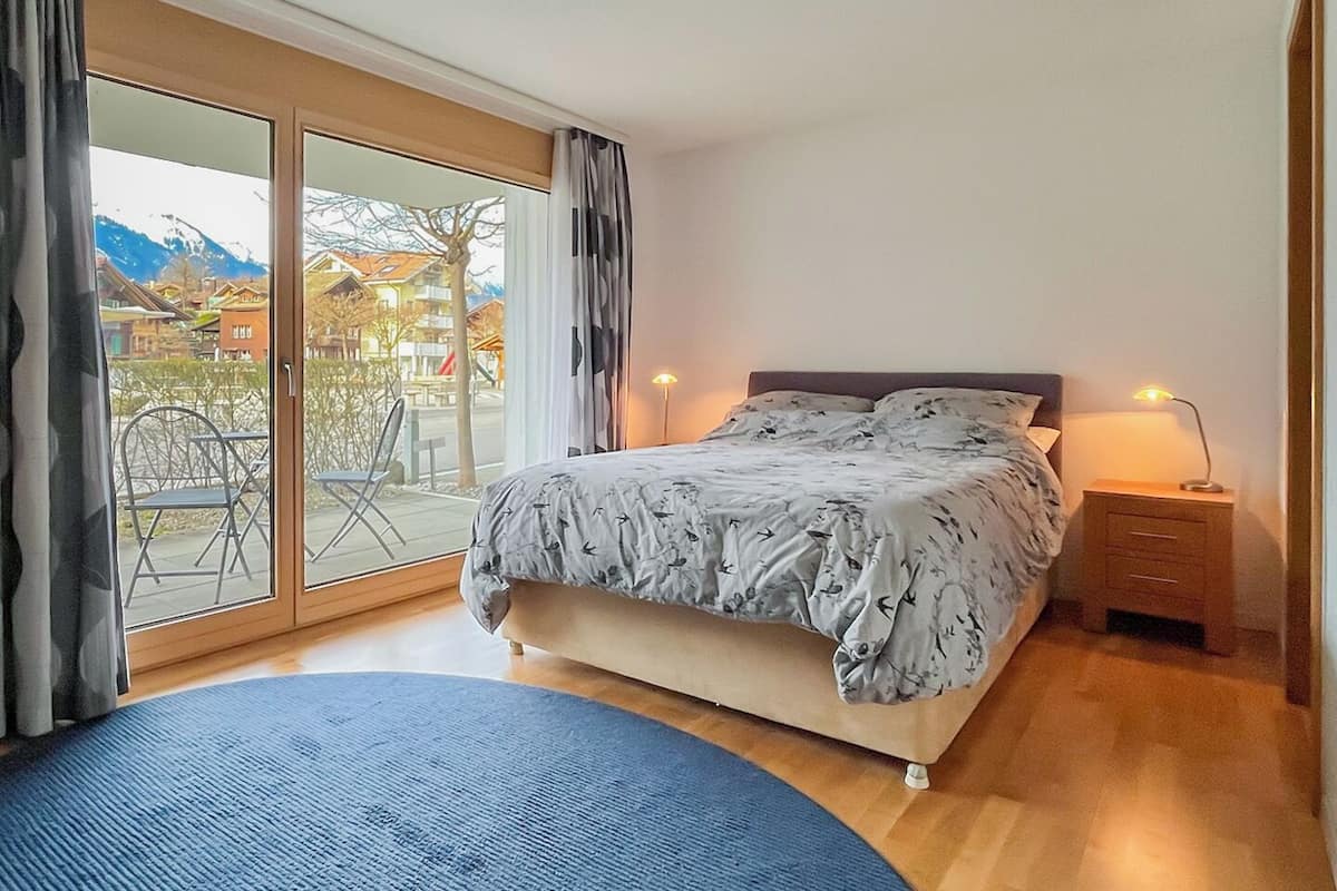 A bedroom with a bed, a nightstand, a lamp, a blue rug, and a sliding glass door leading to an outdoor area with chairs and a table.