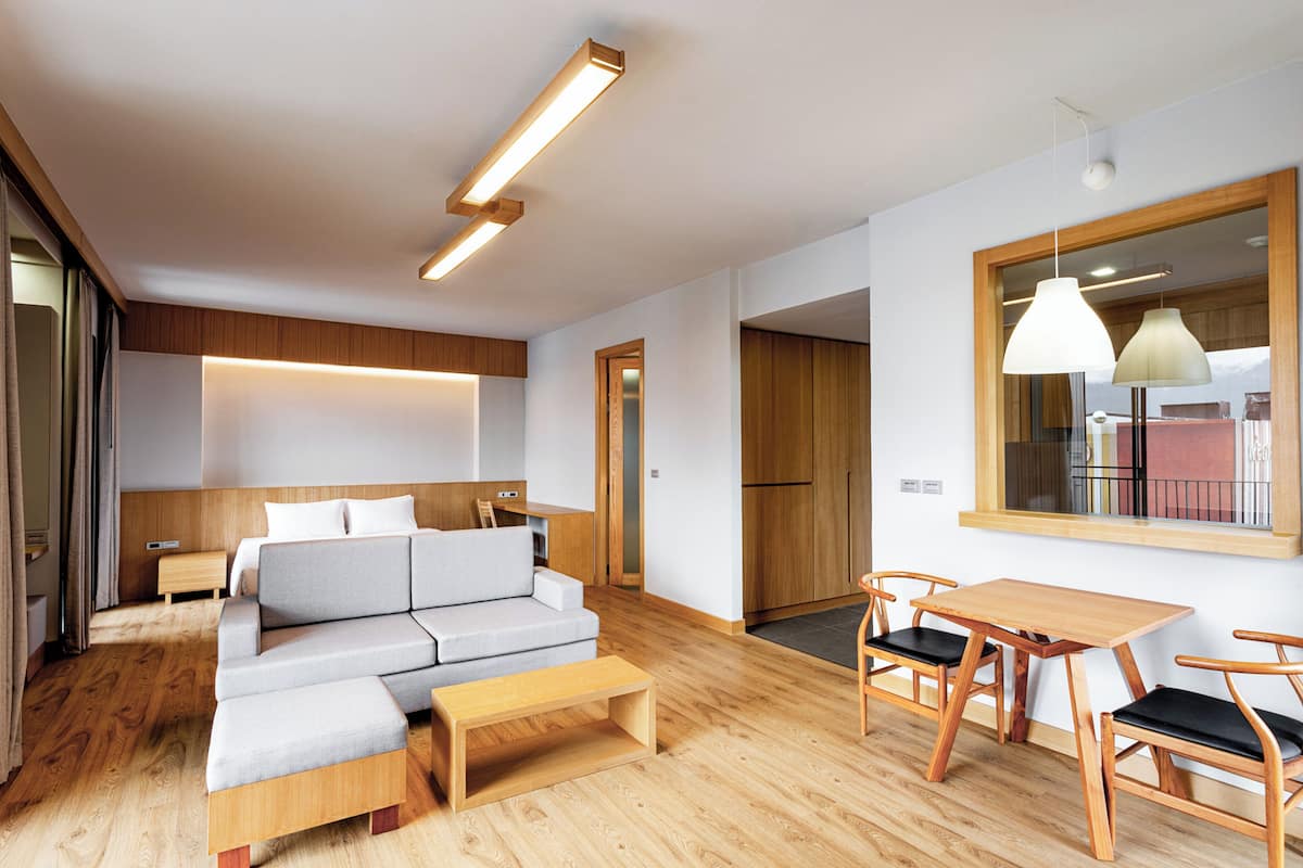 A modern hotel room with a wooden floor, a grey sofa, a wooden dining table with chairs, and a large window with a view.