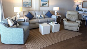 Un salon avec un canapé gris, deux fauteuils, une table basse et un tapis. On y trouve deux lampes, un coussin en forme d’étoile de mer et des tableaux encadrés accrochés au mur.