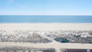 Une plage avec un sentier sablonneux menant à l’eau, bordée d’une végétation clairsemée et d’une clôture en bois.