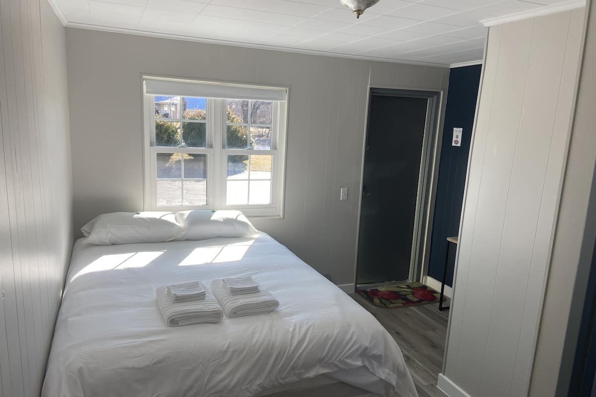 A neatly made bed with white linens and towels, a window with blinds, and a door leading to another room.