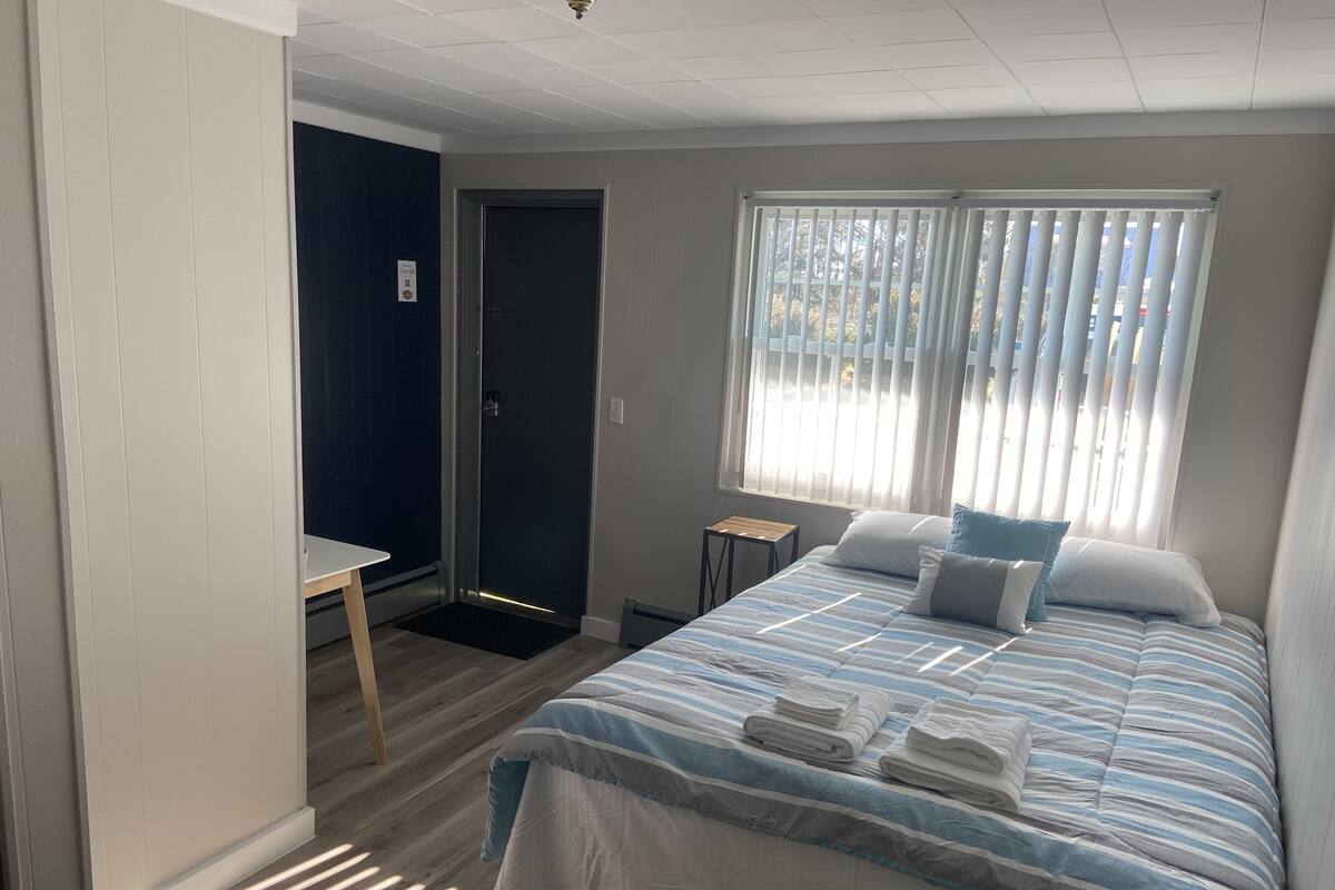A bedroom with a bed, a desk, a door, and a window with blinds.