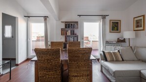 Un salon comprenant une table à manger, quatre chaises en osier, un canapé, une bibliothèque et une fenêtre avec des rideaux.