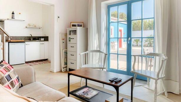 A room with a sofa, a table with a magazine, two white chairs, a kitchen area, and a large window with a view of the outside.