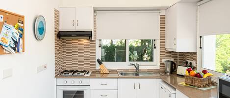 Una cocina moderna con armarios blancos, estufa a gas, horno empotrado, pileta y ventana con vista a follaje.