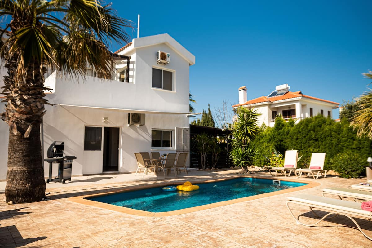 Un espace piscine avec des chaises longues, un barbecue et un bâtiment blanc surmonté d’un toit rouge.