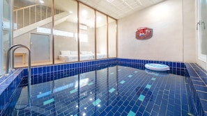 A swimming pool with blue tiles, a glass door, and a bed visible through the glass.