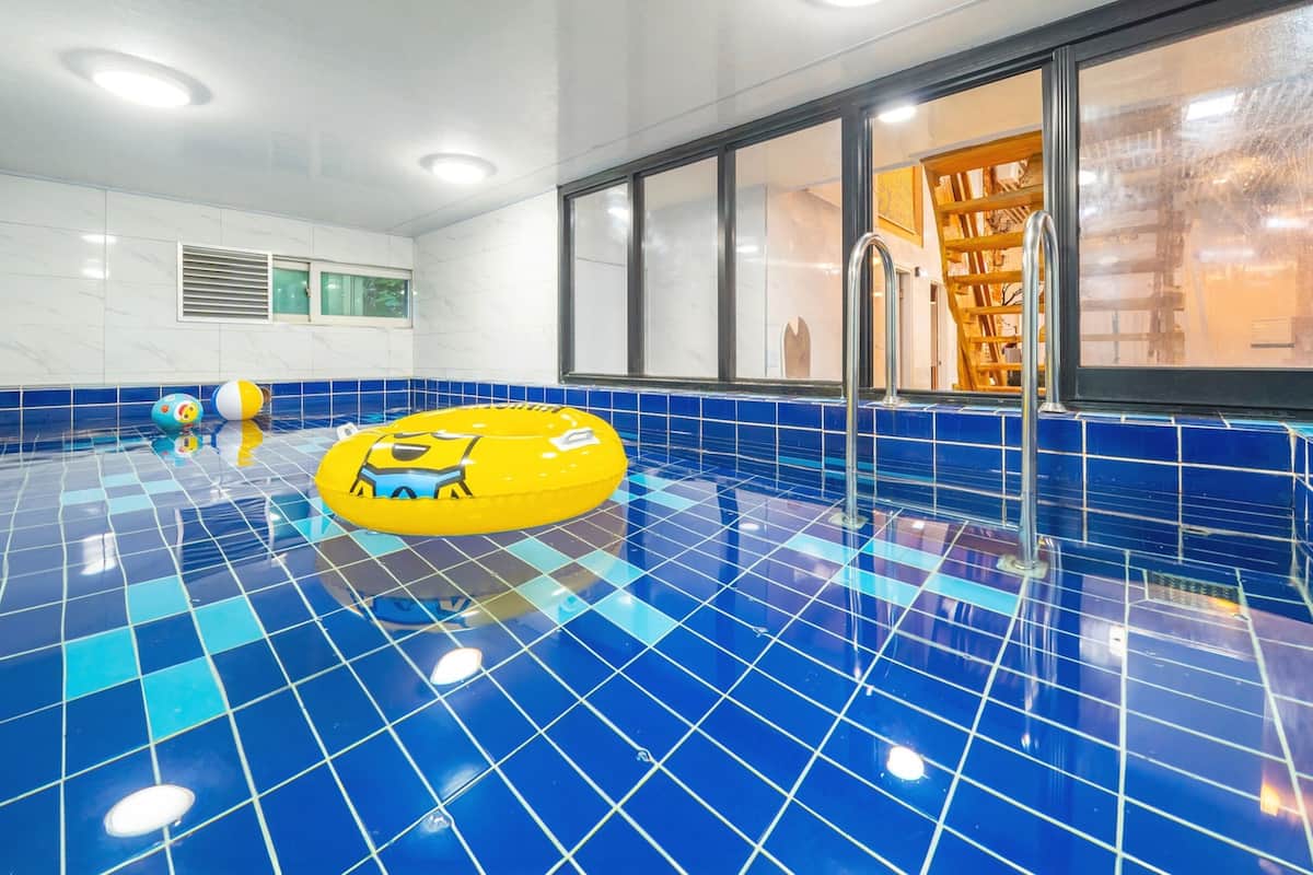A swimming pool with blue tiles, a yellow inflatable ring, and a window with a view of a wooden structure.