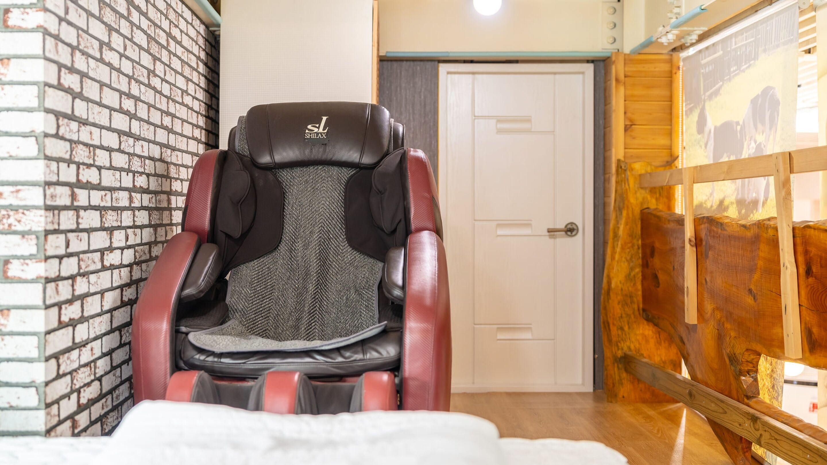 A massage chair with a herringbone-patterned seat and black and red accents, positioned in a room with a brick wall, a wooden panel wall, and a door.
