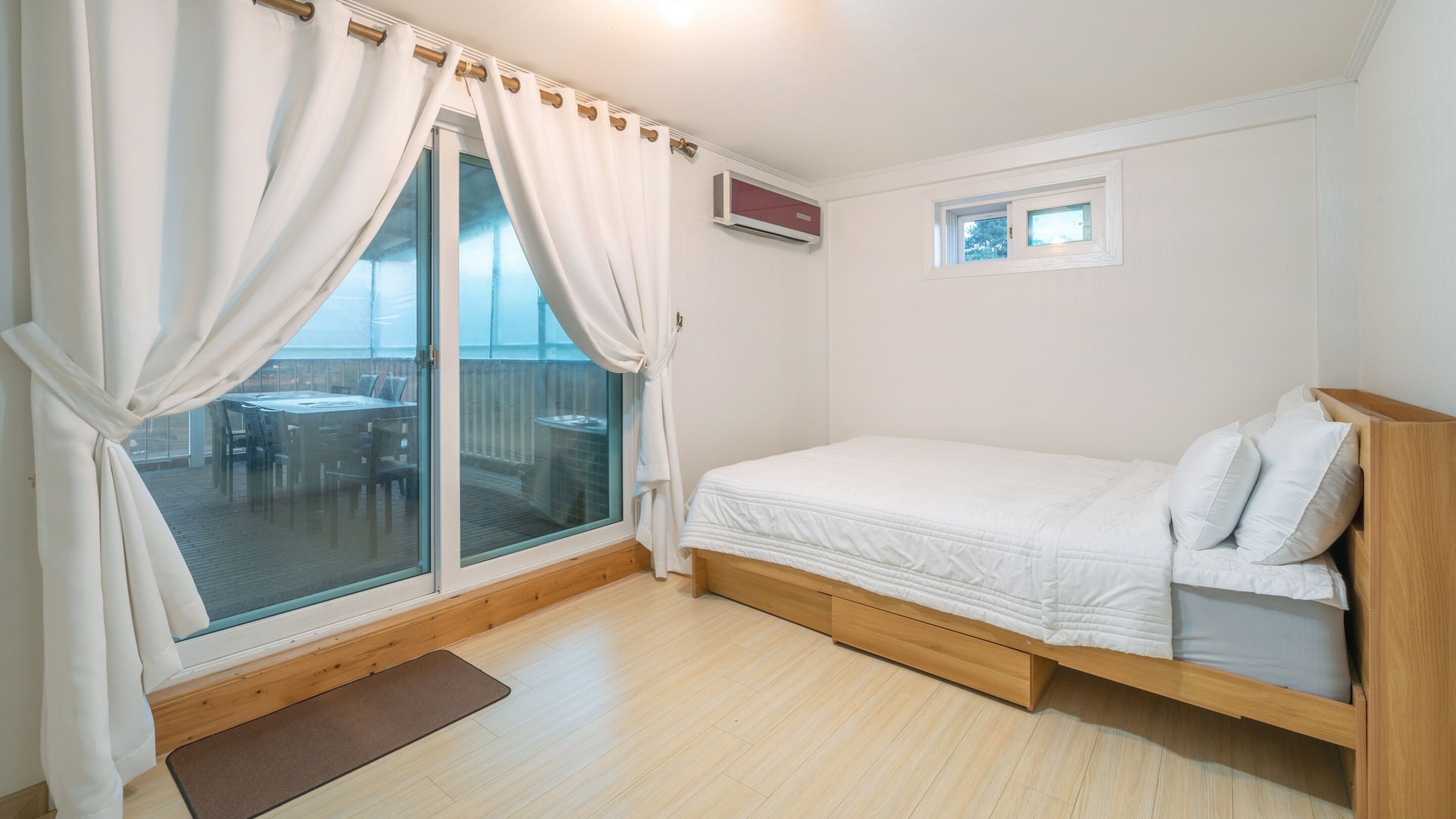 Un dormitorio con cama de madera, una ventana con cortinas, una mesita y vistas a una terraza exterior.