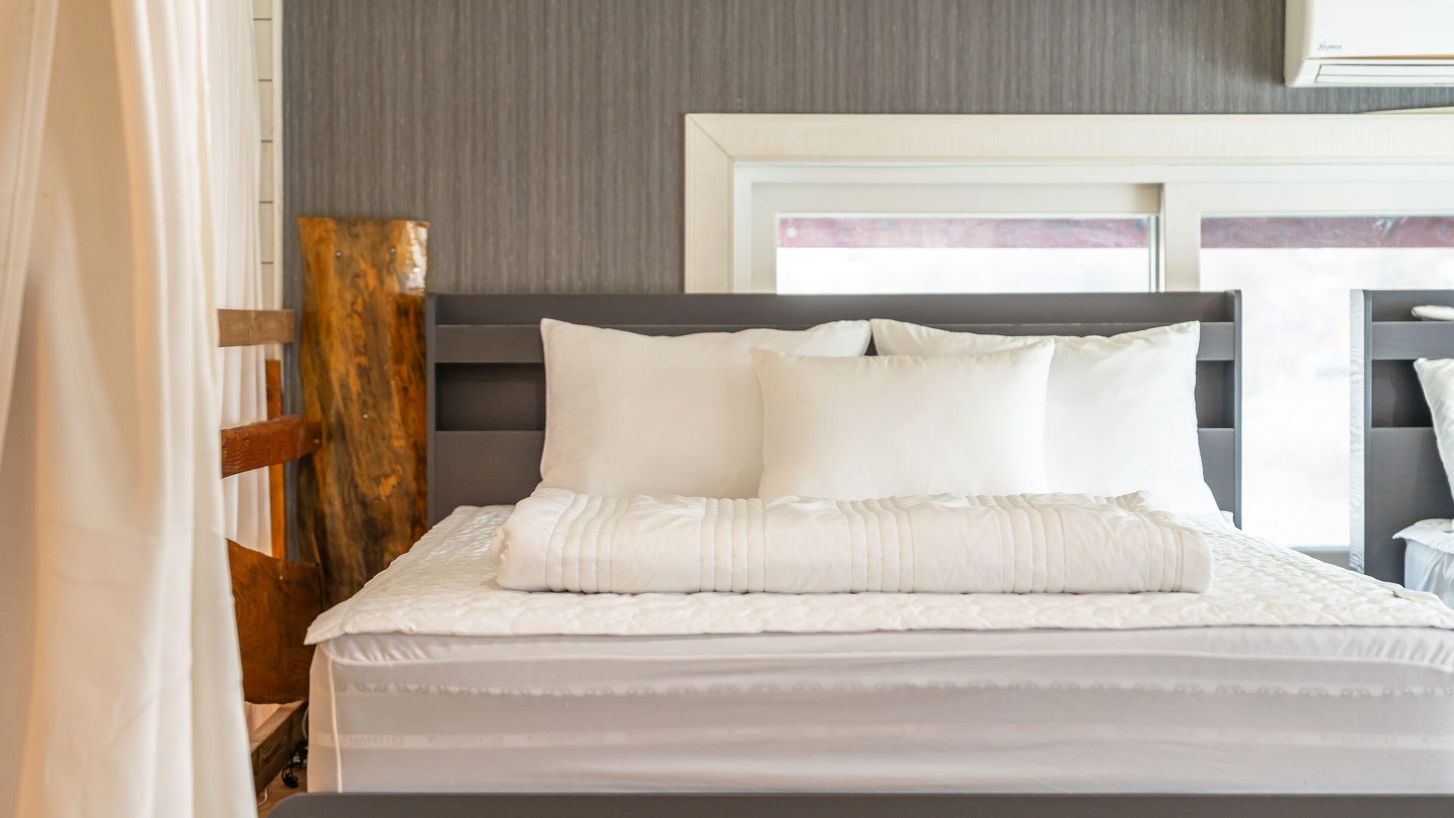 A neatly made bed with white bedding and pillows, a wooden headboard, and a window with a view.