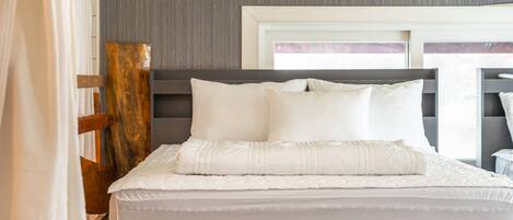 Una cama bien hecha con ropa blanca y almohadas, cabecera de madera, y una ventana con vistas.