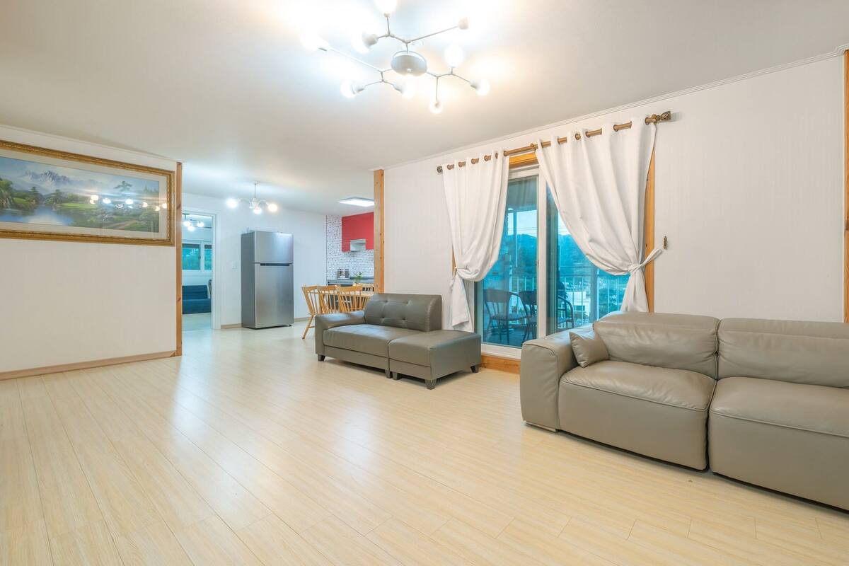 A spacious living area with a grey sofa, a dining table with chairs, a kitchen in the background, and a large window with curtains.