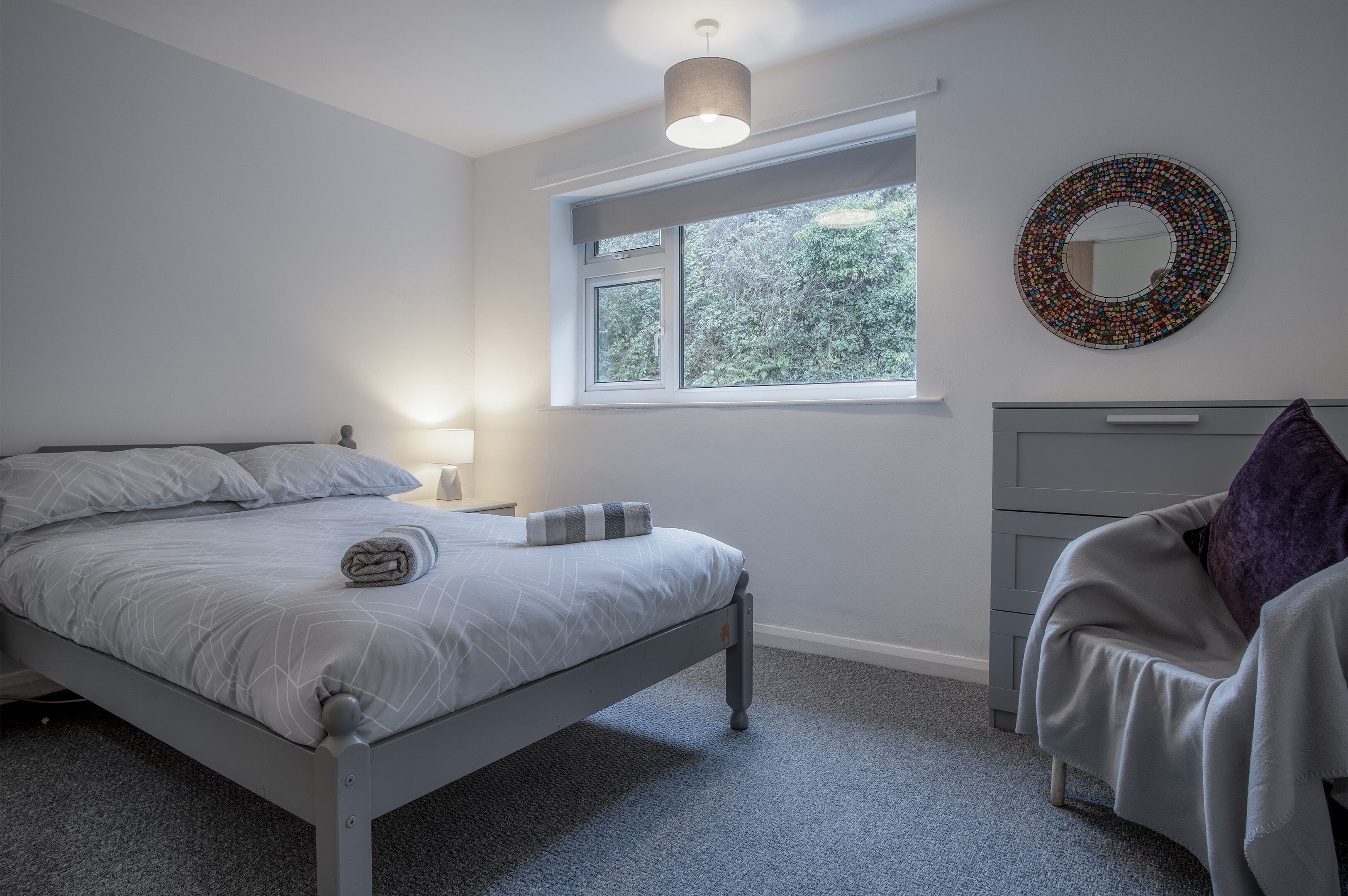 A bedroom with a bed, bedside table, lamp, window, and a chair.