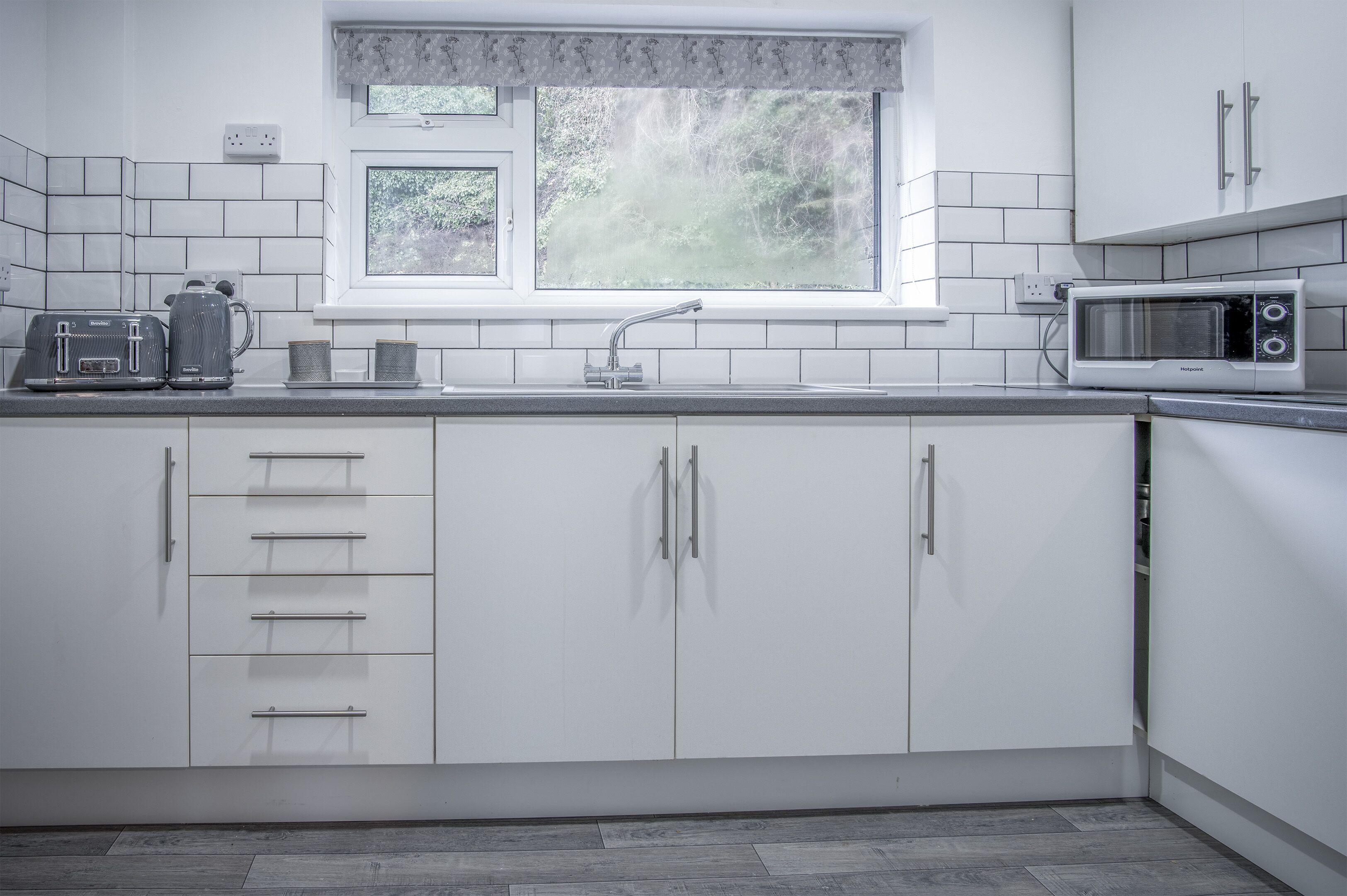A modern kitchen with white cabinets, a window with a view of greenery, a microwave, and a kettle on the counter.
