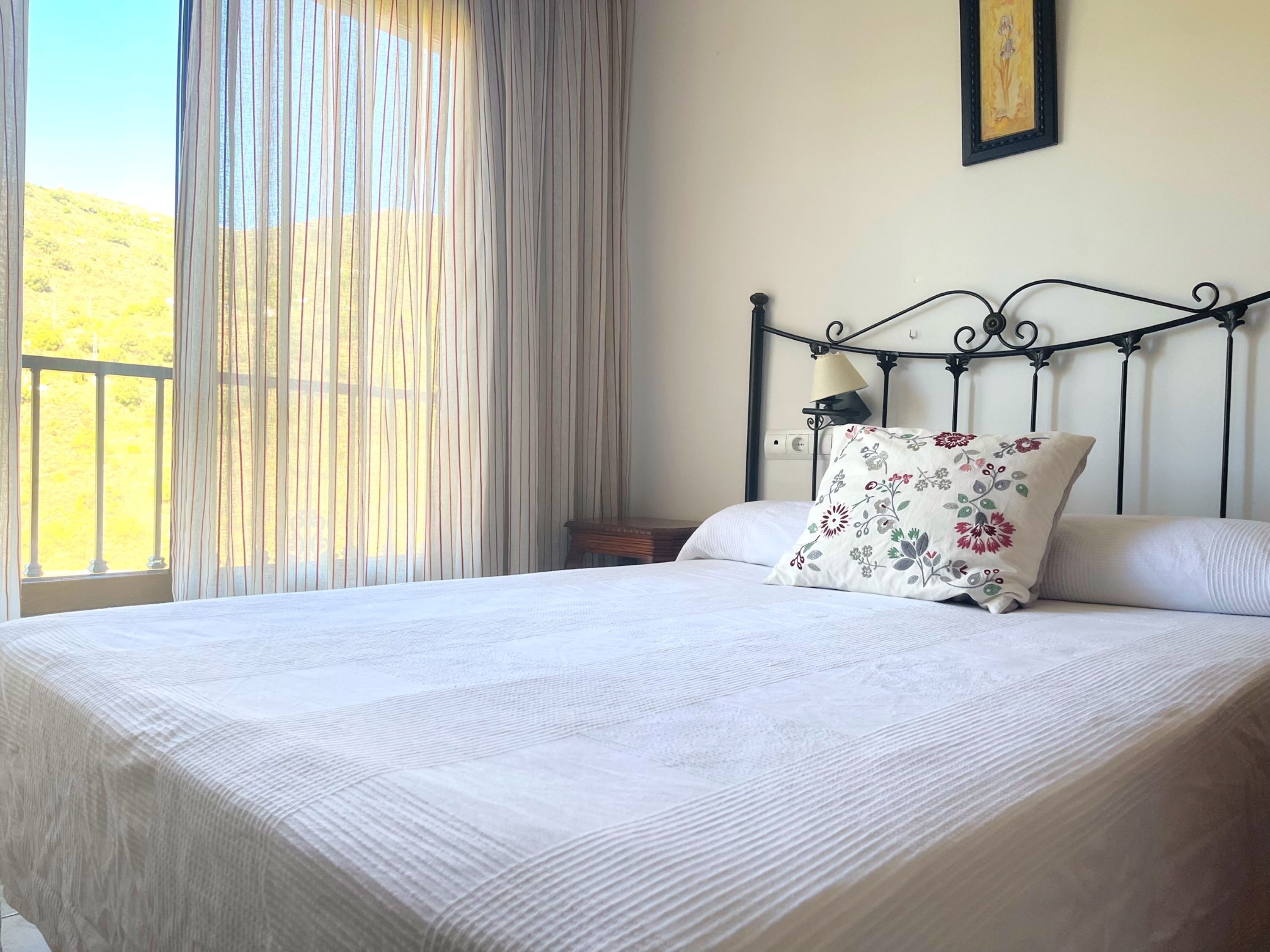 A neatly made bed with a floral pillow, a lamp on a bedside table, and a view of greenery outside.
