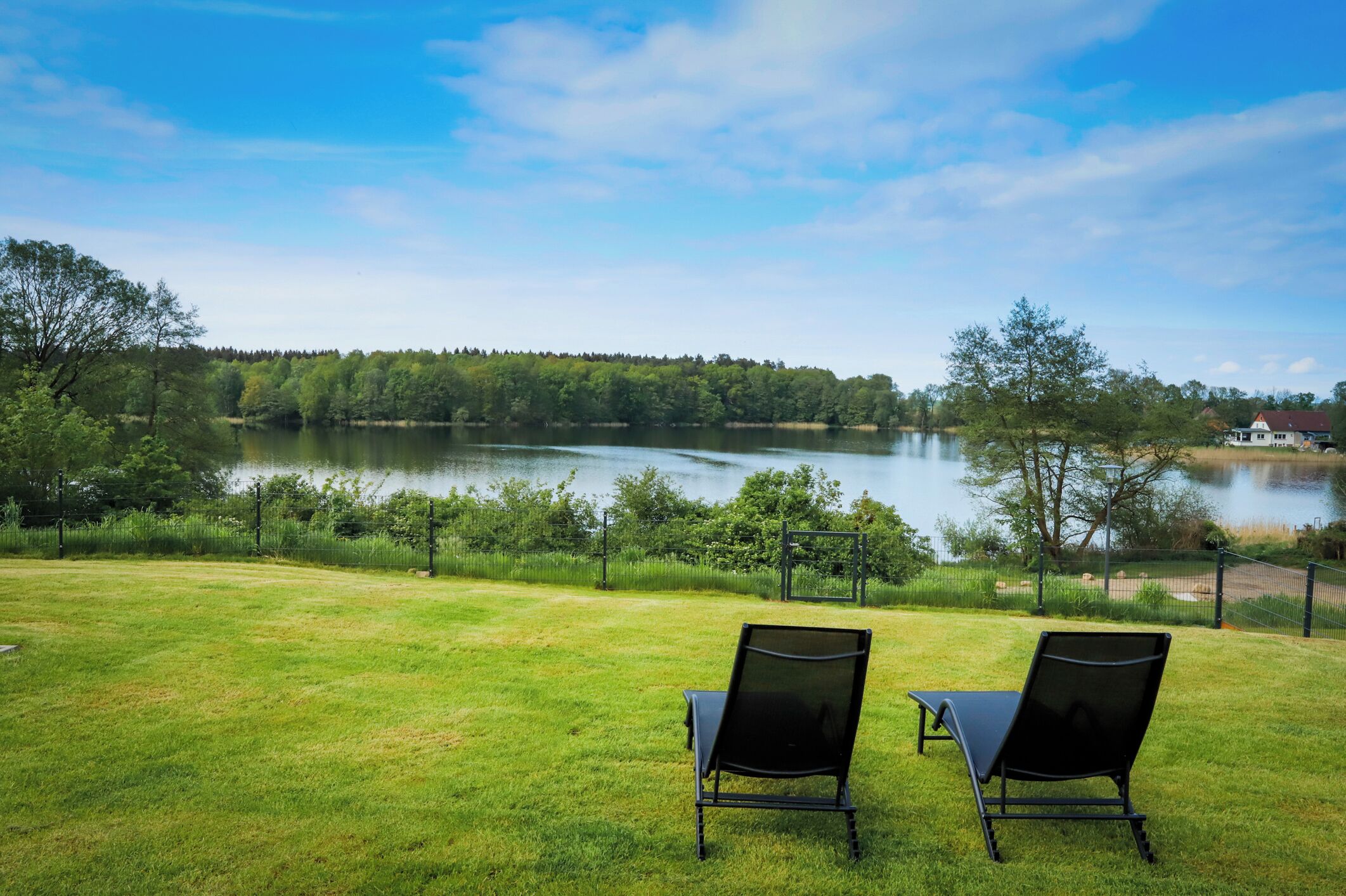 A grassy area with two black chairs facing a lake, surrounded by trees and a fence.