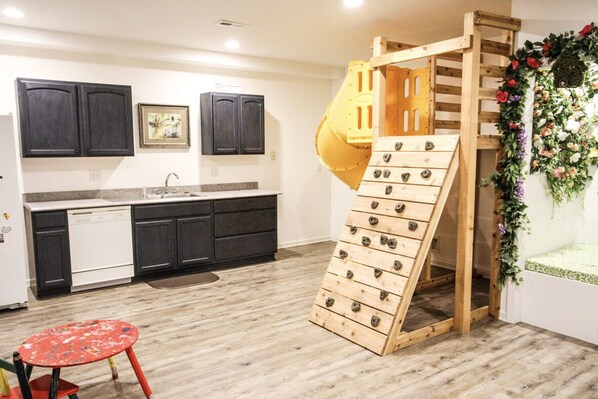 Una cocina con armarios oscuros, lavavajillas, un parque infantil de madera con tobogán y una mesa roja.