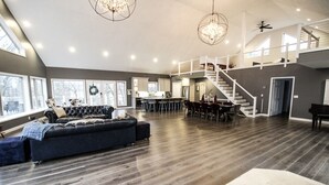 Amplia sala de estar con piso de madera oscura, un sofá grande y oscuro, una mesa de comedor con sillas y una escalera que lleva a un nivel superior.